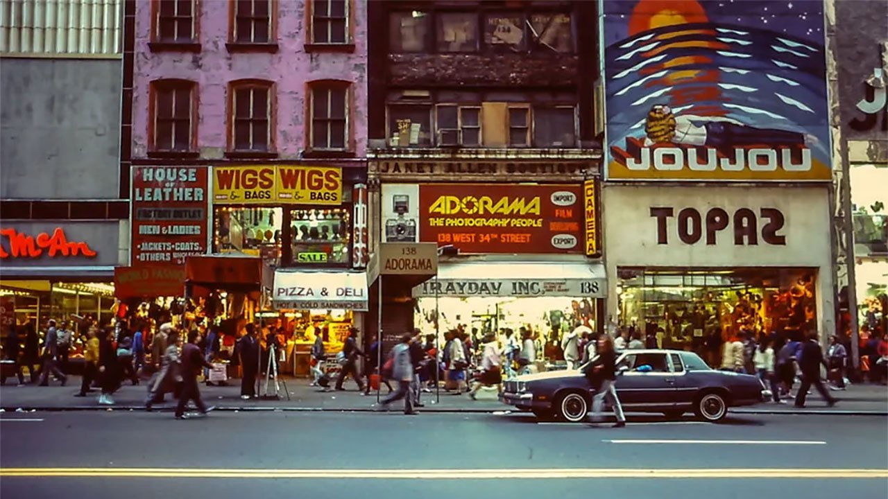 Rušná pouliční scéna v New Yorku s chodci, obchody, zaparkovaným autem a barevným značením se zaměřením na umístění výlohy Adorama.