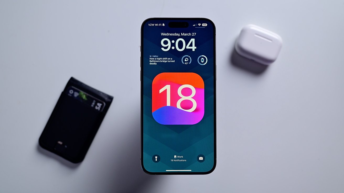 iPhone se zamykací obrazovkou s ikonou iOS 18.  Nedaleko je peněženka a pouzdro na AirPods.