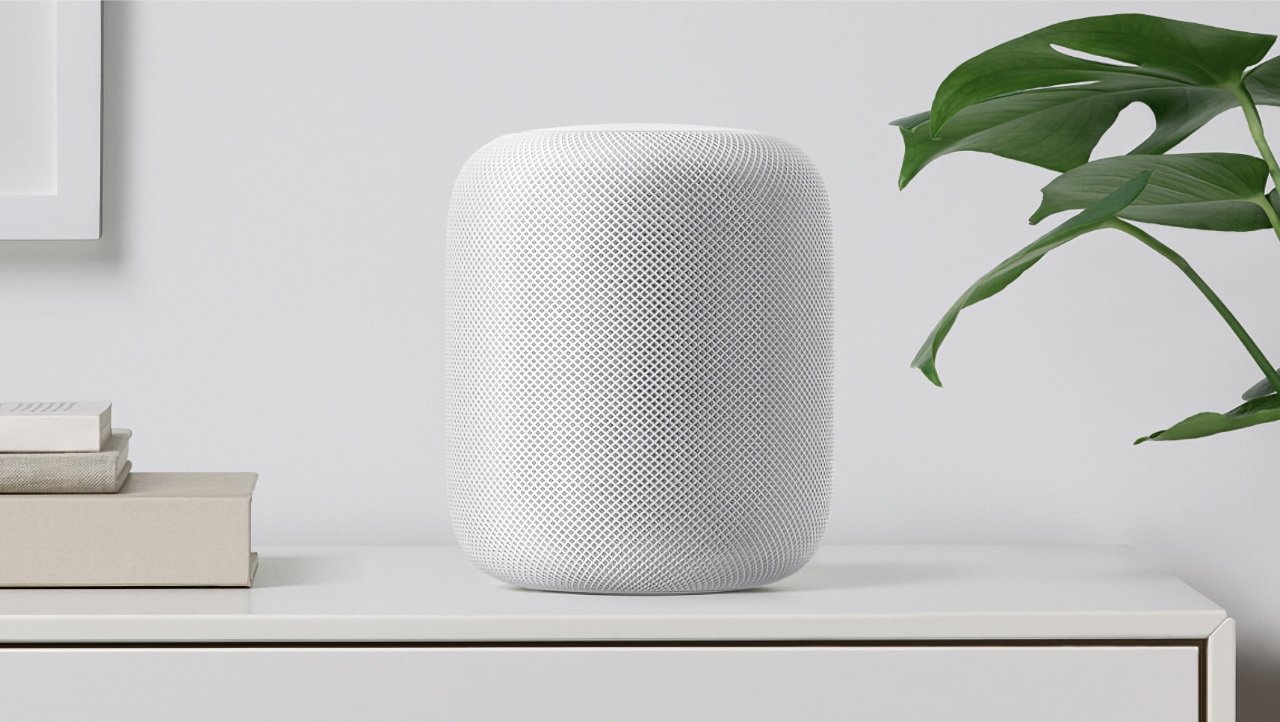 Bílý válcový reproduktor HomePod na polici s rostlinou a knihami v minimalistickém pokoji.