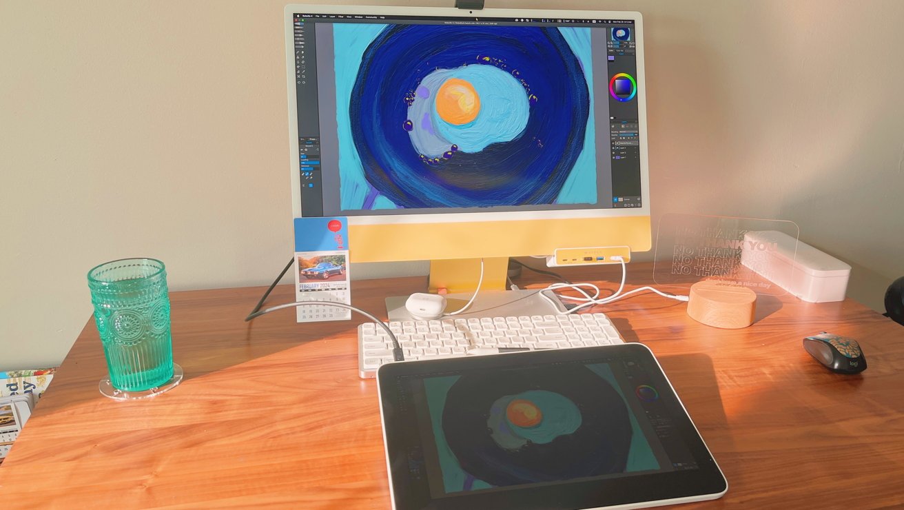 Dobře osvětlený pracovní prostor s monitorem zobrazujícím grafiku, tablet, klávesnice, myš, sklenice vody, kalendář