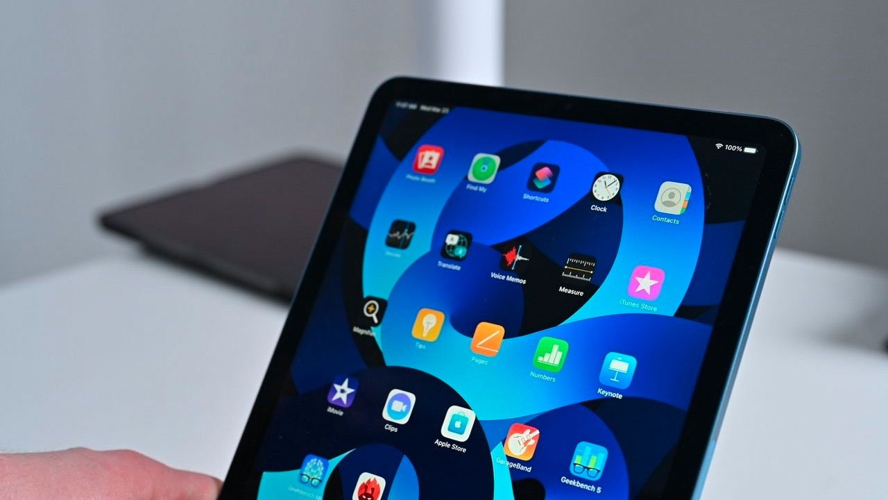 Ruka držící tablet iPad Air zobrazující barevné ikony aplikací na obrazovce s rozmazaným pozadím.
