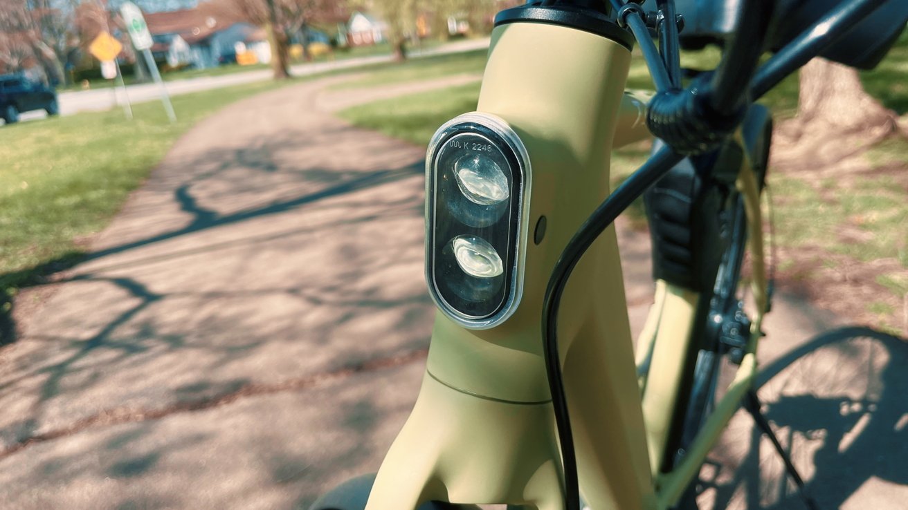 Detail hlavové trubky a vidlice jízdního kola s připojeným dvojitým LED světlem na rozmazaném pozadí parkové cesty.
