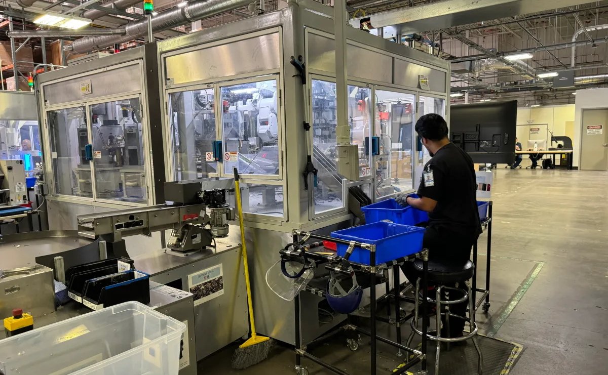 Dělník v továrně obsluhuje stroj poblíž modrých popelnic a různého vybavení v prostorném, dobře osvětleném průmyslovém prostředí s viditelným potrubím a bezpečnostními značkami.