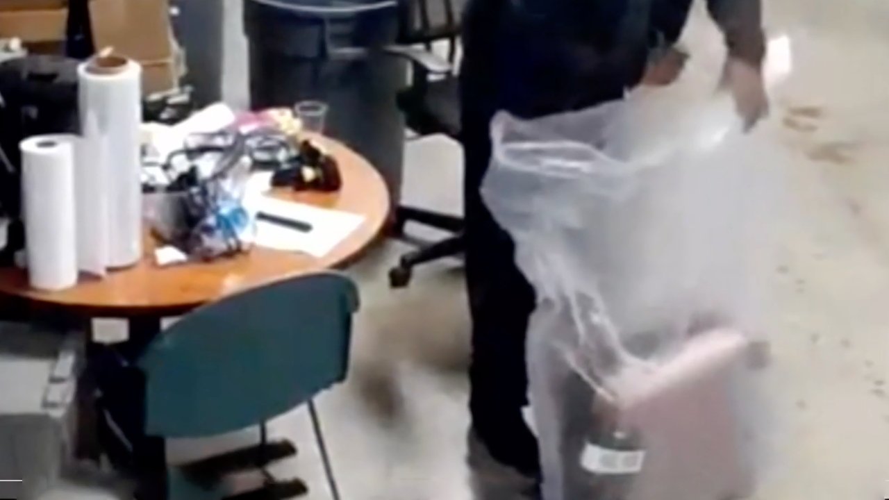 Osoba v kancelářském prostředí balící předmět do plastu.  Nedaleko je zaneřáděný kulatý stůl se židlemi, rolemi papíru a kancelářskými potřebami.