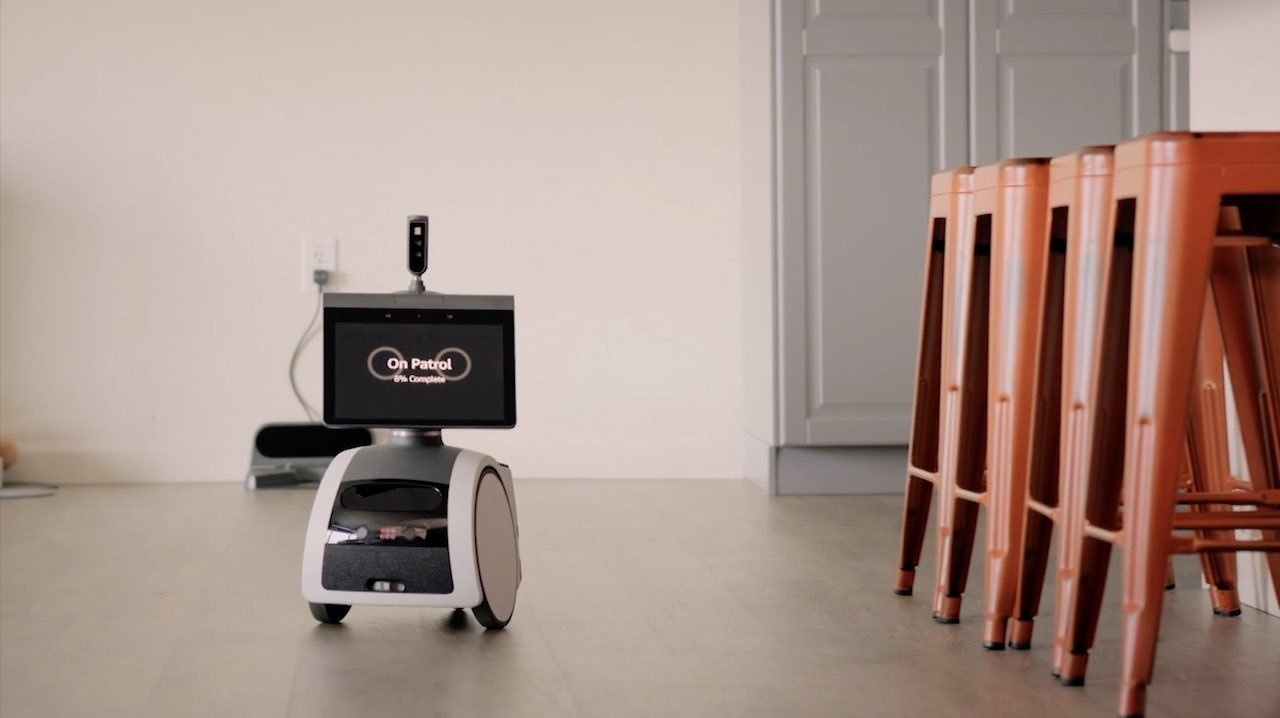 Malý Astro robot s displejem „On Patrol“ stojí na podlaze poblíž oranžových židlí a šedých skříní.